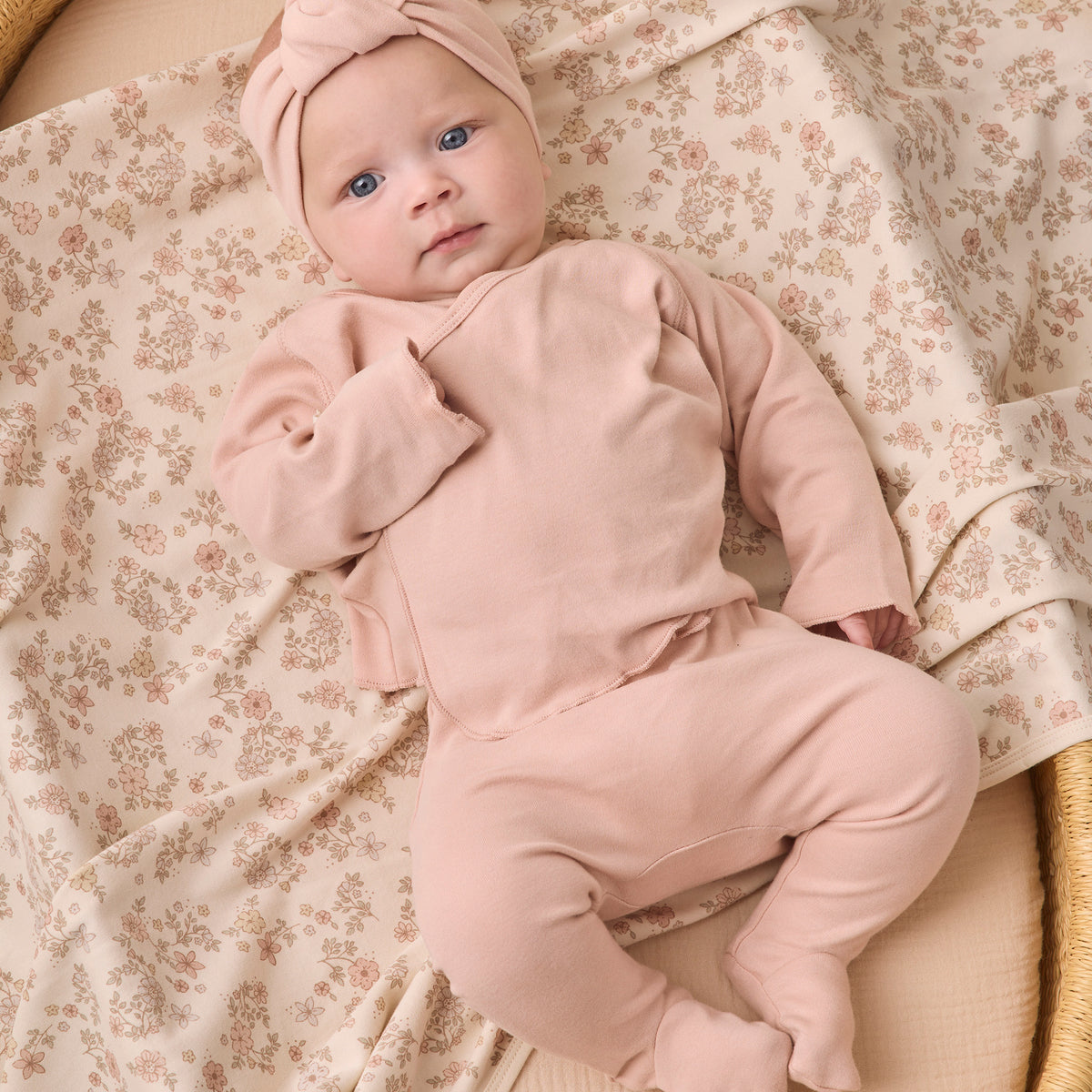 A baby lying on a woven bassinet, wearing a dusty pink footed sleeper and matching knotted headband, with a cream floral blanket underneath.