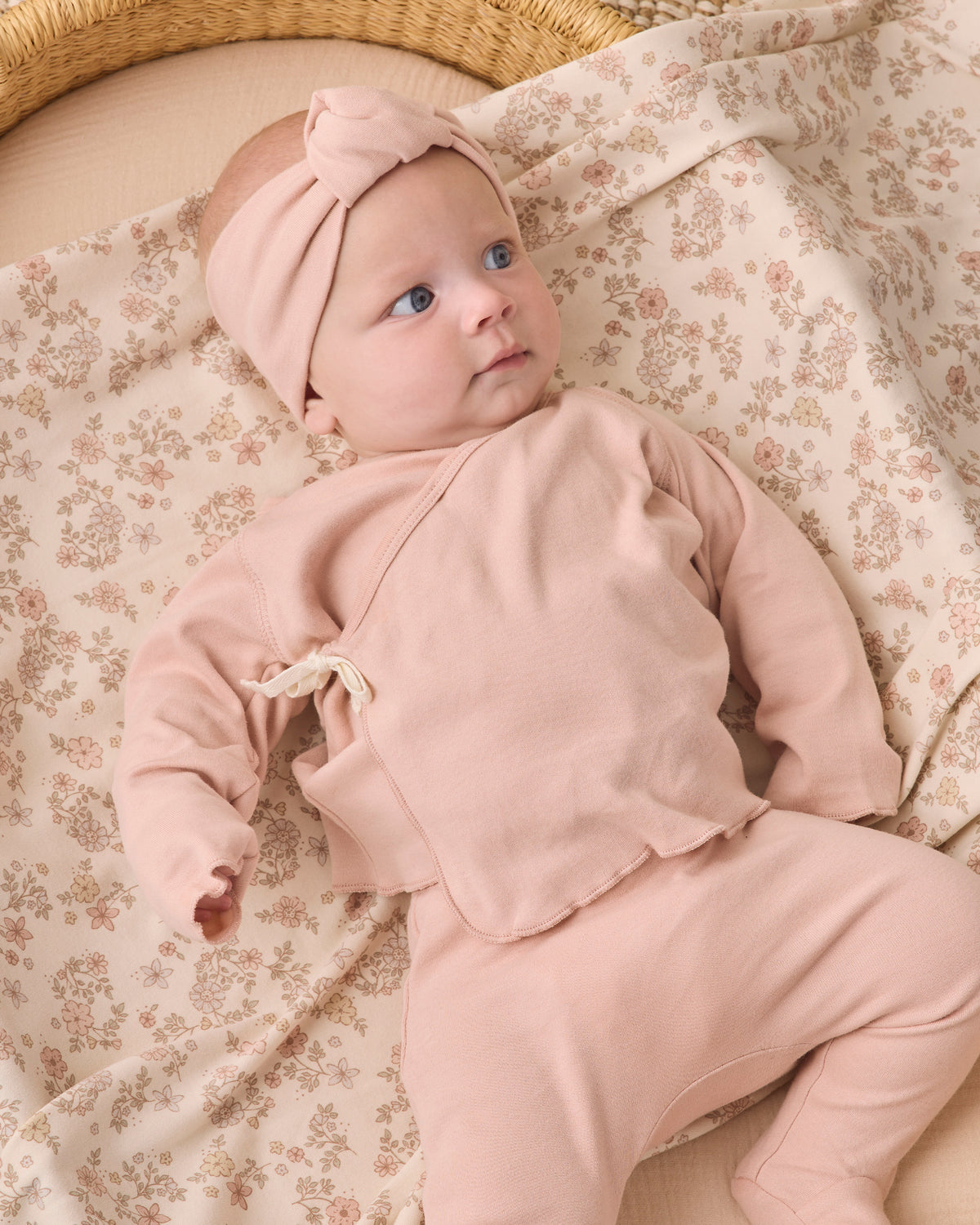 A baby lying on a floral blanket wearing a matching dusty pink two-piece outfit with a knotted headband.