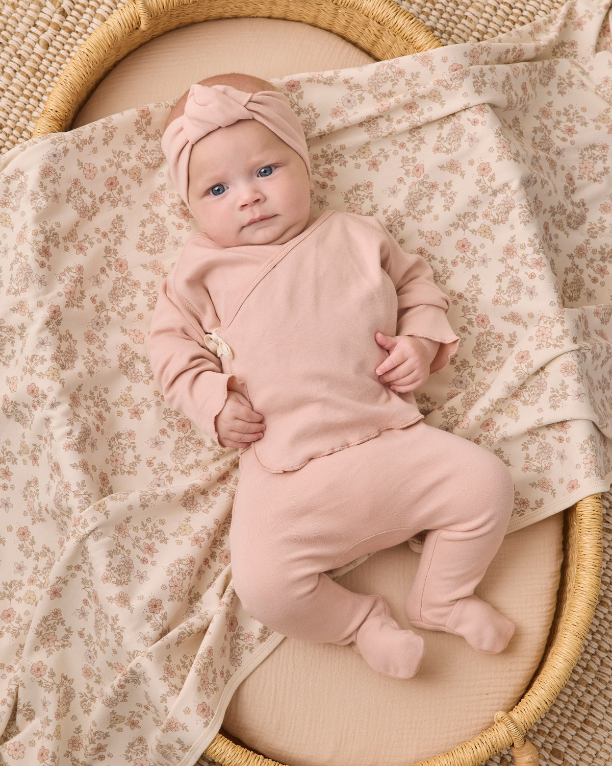 A baby wearing a dusty pink footed sleeper and matching bow headband, lying on a floral-patterned blanket inside a woven basket.