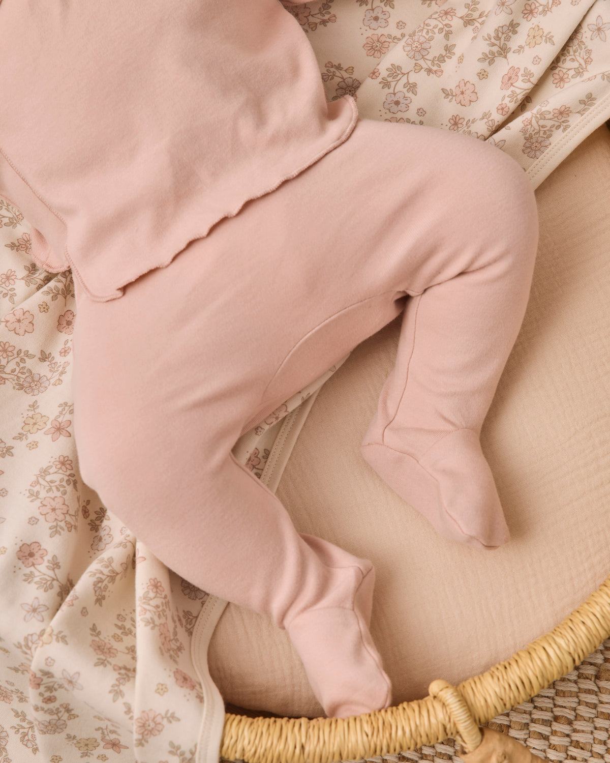 A close-up of a baby wearing dusty pink footed leggings and a matching top, lying on a cream-colored woven bassinet with floral-patterned blankets.