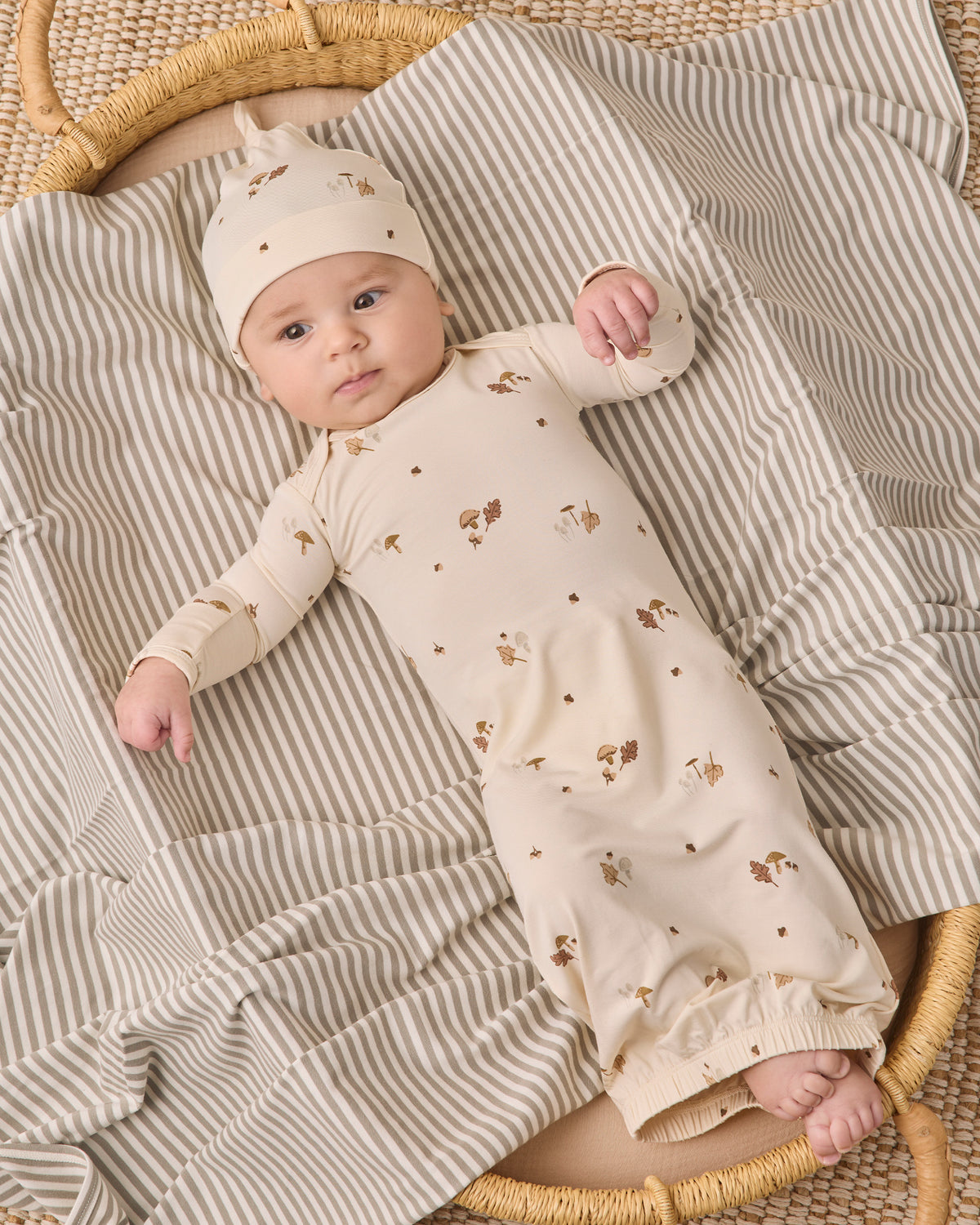 A baby lying on a striped blanket in a woven basket, wearing a cream long-sleeve romper with small brown animal prints and a matching beanie.