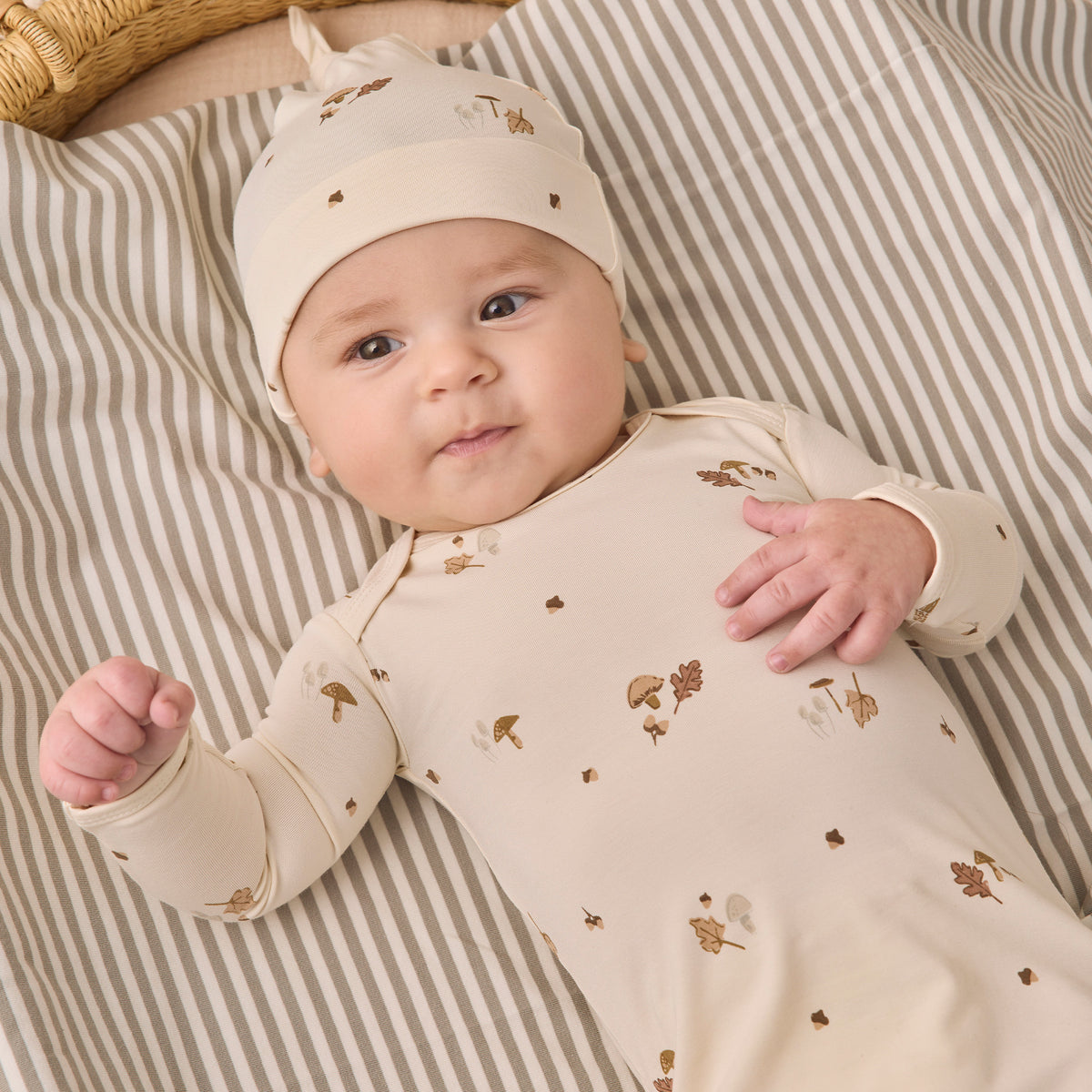 A baby lying on a striped blanket next to a woven basket, wearing a cream-colored long-sleeve bodysuit and matching beanie hat with small brown and gold woodland prints.