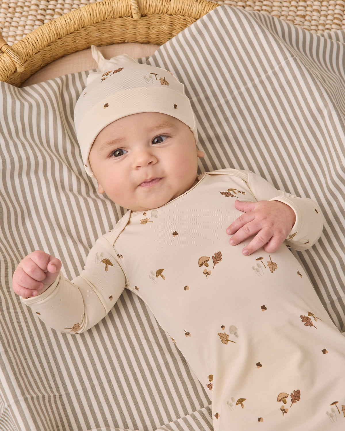 A baby lying on a striped blanket next to a woven basket, wearing a cream-colored long-sleeve bodysuit and matching beanie hat with small brown and gold woodland prints.