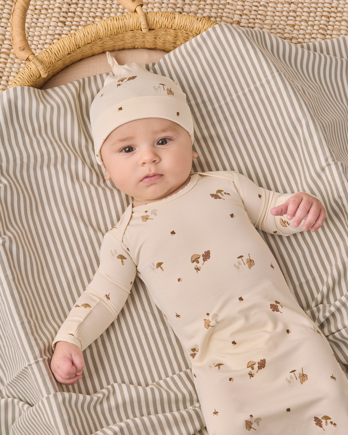 A baby lying on a striped blanket next to a woven basket, wearing a cream-colored long-sleeve bodysuit and matching hat with small brown animal prints.
