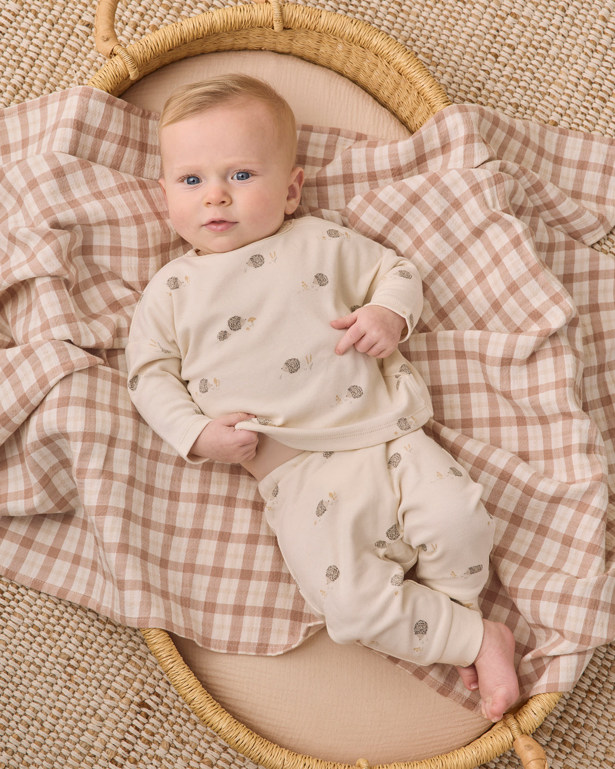 A baby lying on a woven mat, wearing a cream long-sleeve romper and matching pants with small gray tree prints, while resting on a beige and brown gingham blanket inside a wicker basket.