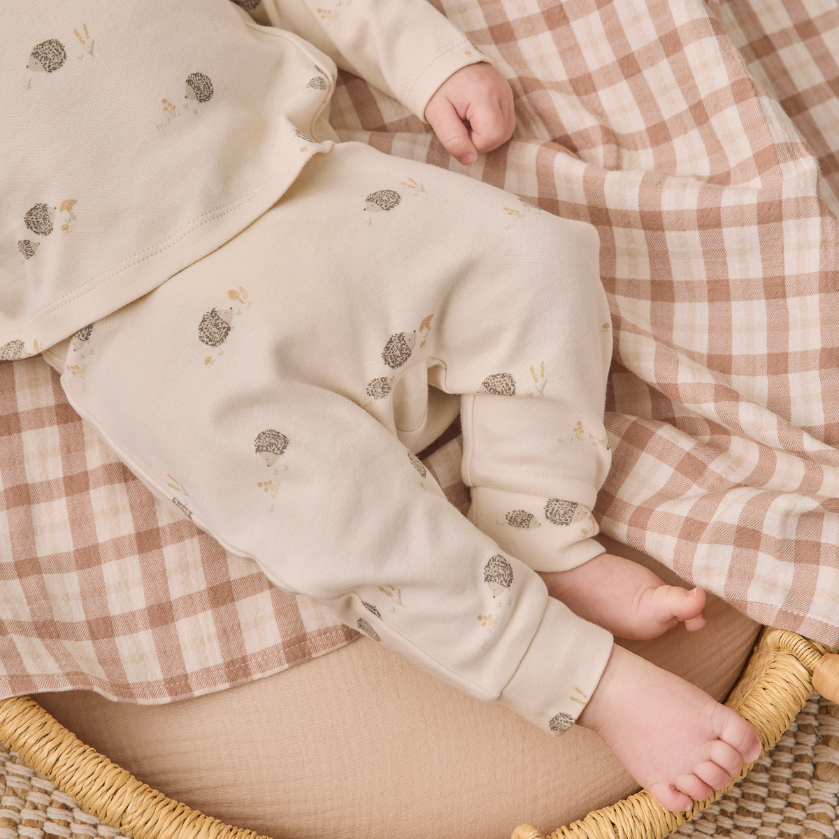 A baby wearing a cream-colored long-sleeve romper and matching pants with small gray and beige floral prints, lying on a brown and white checkered blanket inside a woven rattan bassinet.