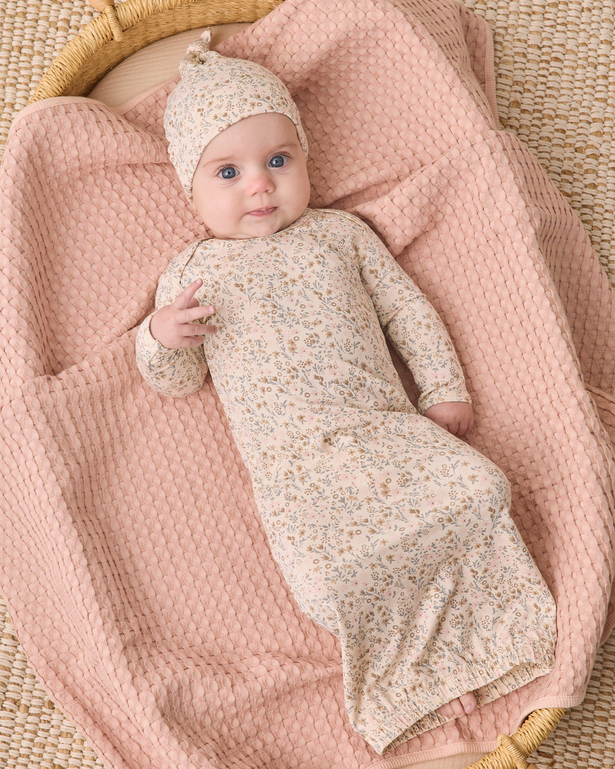 A baby wearing a cream long-sleeve swaddle blanket and matching beanie hat with a delicate floral print, lying on a pink textured blanket inside a woven basket.