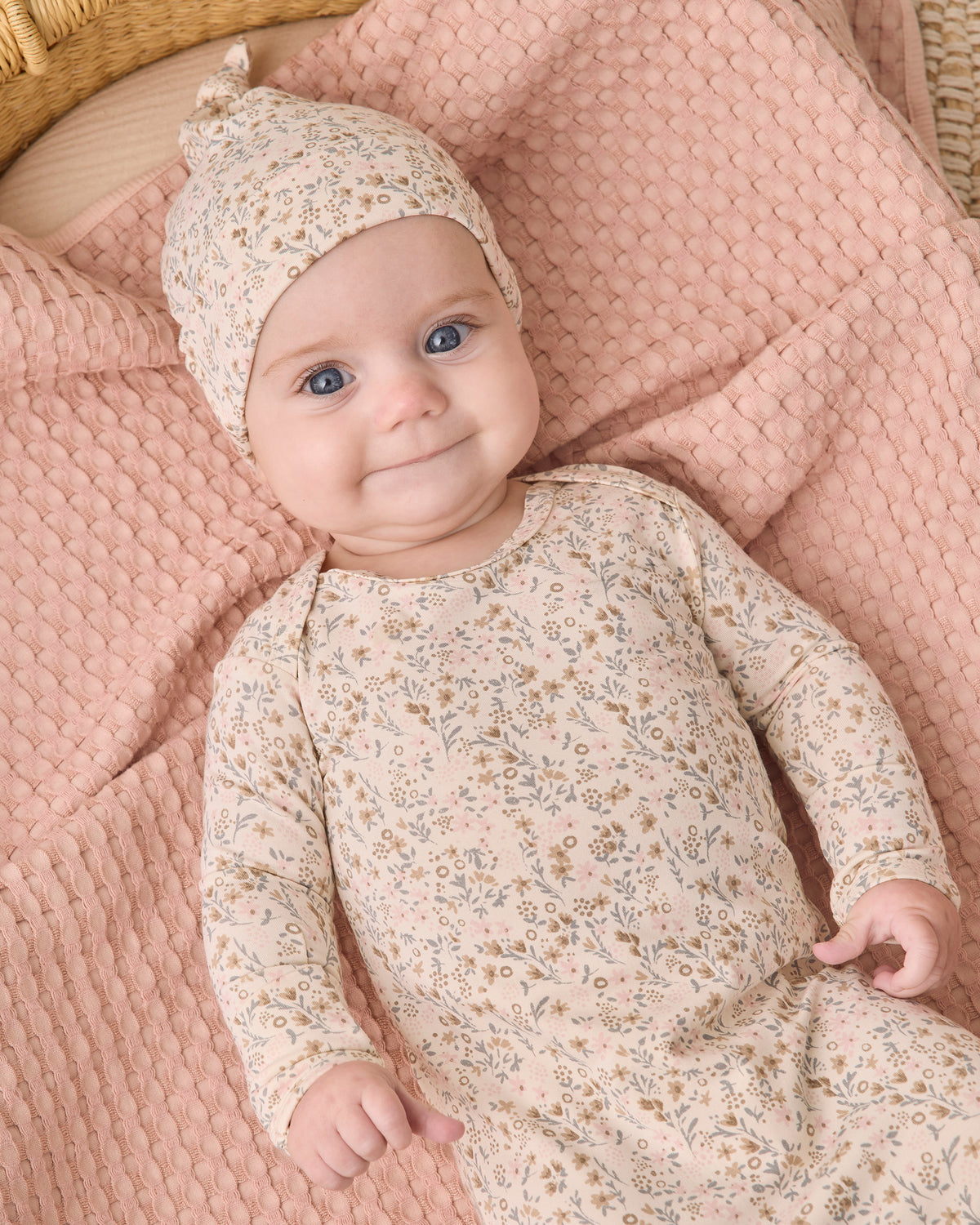 A baby lying on a pink textured blanket, wearing a matching long-sleeve romper and bonnet with a small beige and brown floral print.