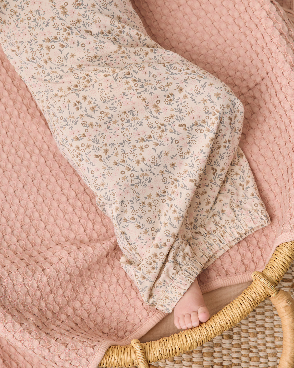 A close-up of a baby blanket with a small beige and blue floral print, draped over a textured dusty pink waffle-knit blanket on a woven rattan chair, with a baby's foot visible.