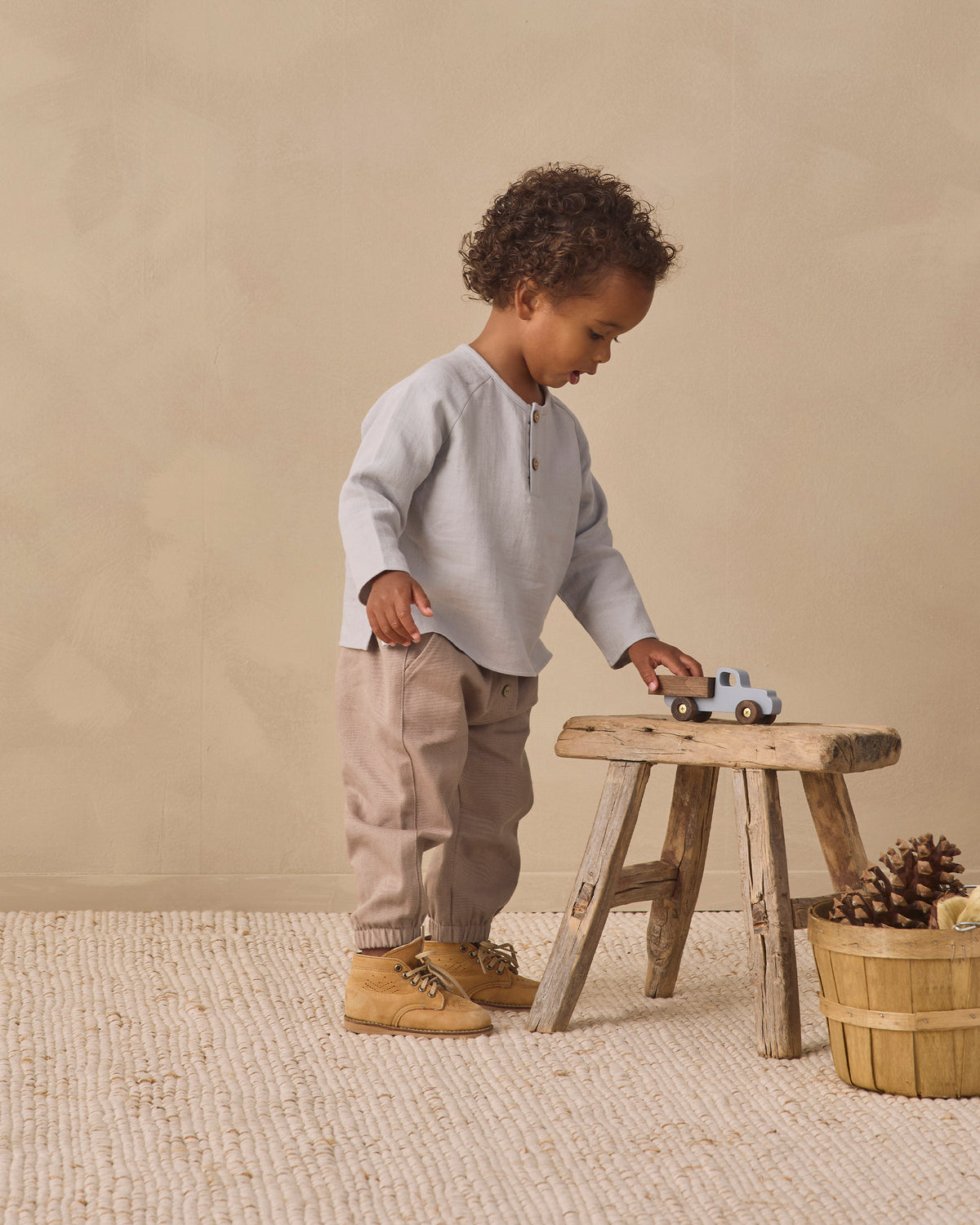A toddler stands beside a small wooden stool in a neutral studio setting, wearing a light gray long-sleeve henley shirt, beige pants, and tan boots, while touching a toy truck on the stool.