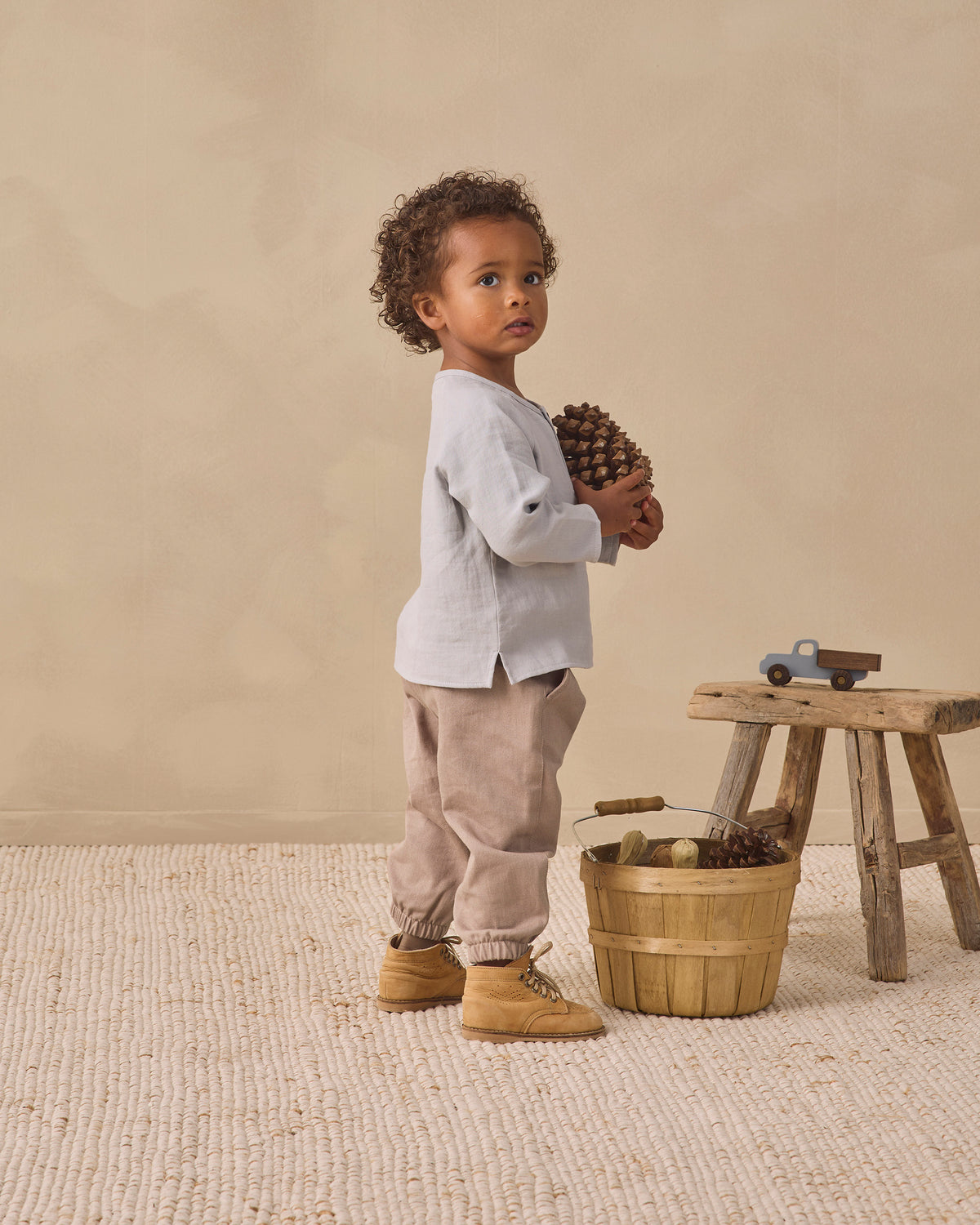 A toddler stands in a rustic studio setting wearing a light gray long-sleeve top, beige pants, and tan boots, holding a pinecone next to a small wooden stool and basket.