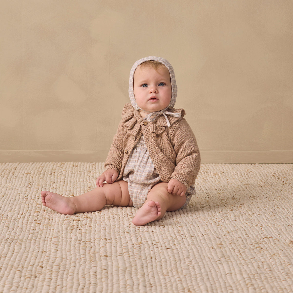 A baby sitting on a textured cream woven rug against a warm beige studio wall, wearing a beige knit bonnet and matching knit outfit.
