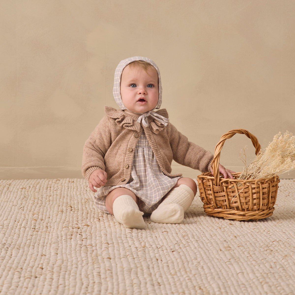 A baby wearing a beige knit bonnet, a brown ruffled cardigan, and a cream plaid romper sits on a textured cream rug next to a small wicker basket with dried grass.