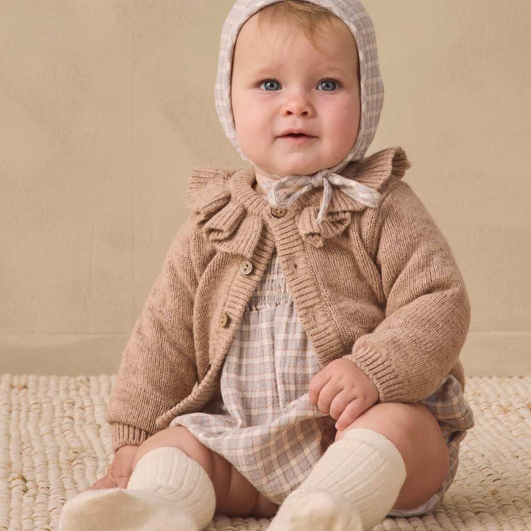 A baby sitting on a textured cream rug against a warm beige wall, wearing a beige knit bonnet, a brown cardigan, and cream socks, with a wicker basket to the side.
