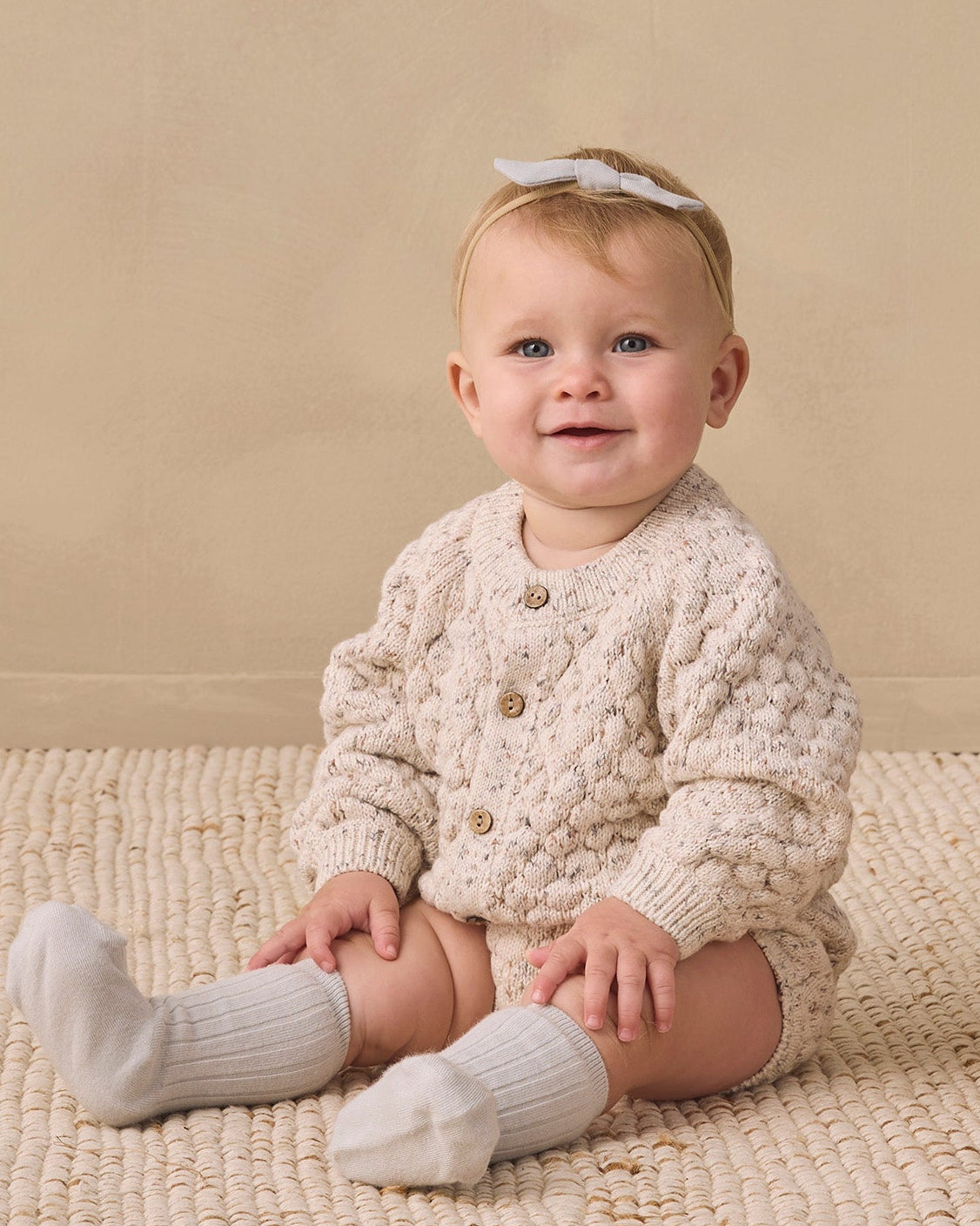 A baby sitting on a woven rug wearing a cream cable-knit cardigan with buttons, matching cream knit socks, and a white bow headband, set against a warm beige background.