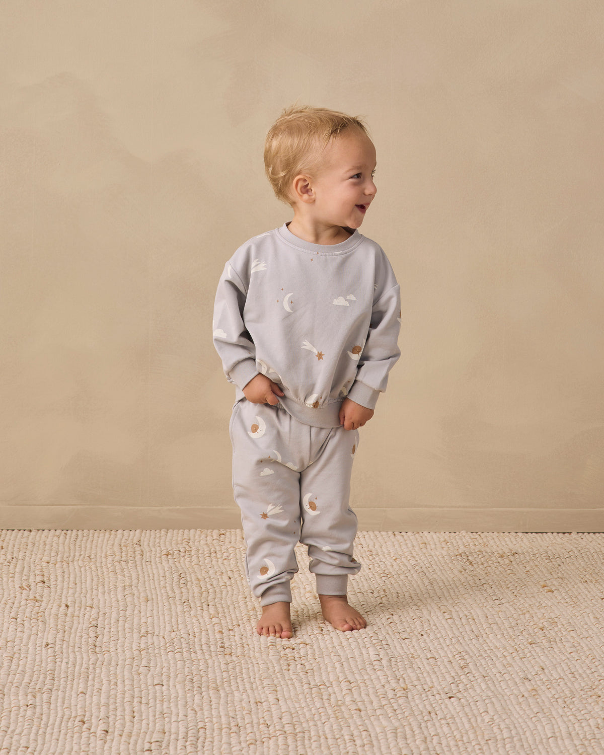 A toddler standing barefoot on a textured cream rug against a warm beige wall, wearing a light gray long-sleeve sweatshirt and matching sweatpants with a small celestial print of stars and crescent moons.