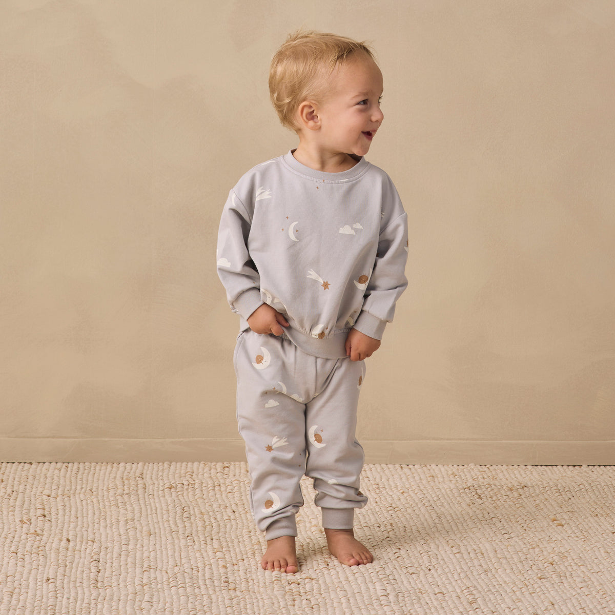 A toddler standing barefoot on a textured cream rug against a warm beige wall, wearing a light gray long-sleeve sweatshirt and matching sweatpants with a small celestial print of stars and crescent moons.