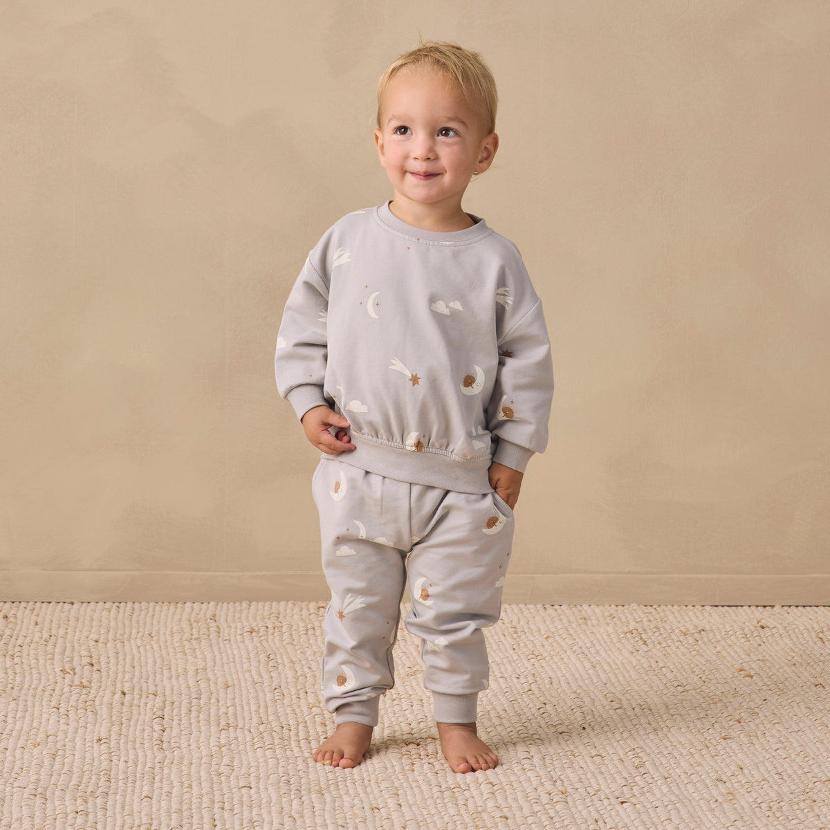 A toddler stands barefoot on a textured cream rug against a warm beige studio wall, wearing a light gray long-sleeve sweatshirt and matching jogger pants with small scattered animal illustrations.