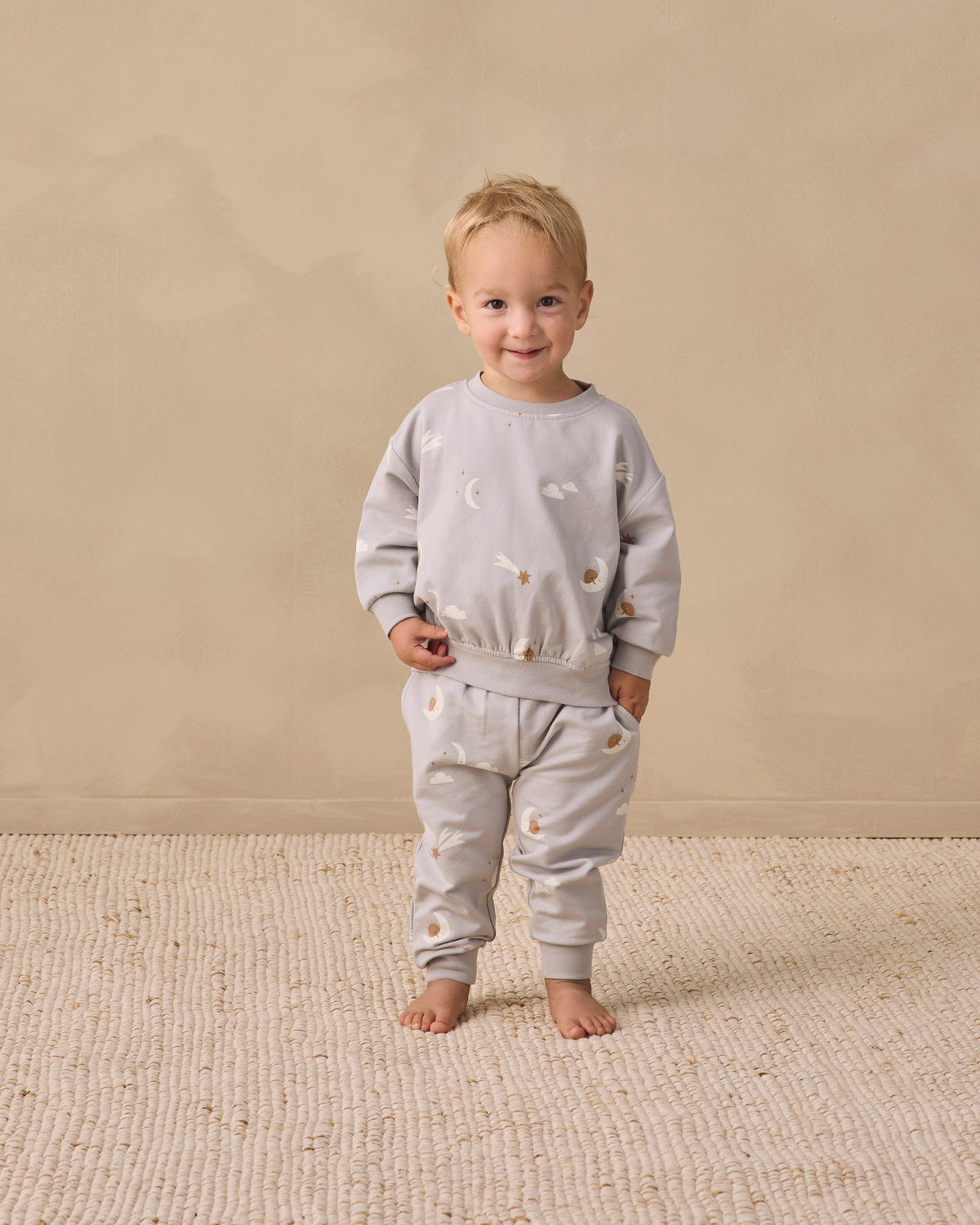 A toddler stands barefoot on a textured cream rug against a warm beige wall, wearing a matching light gray sweatshirt and jogger pants set with small embroidered details.