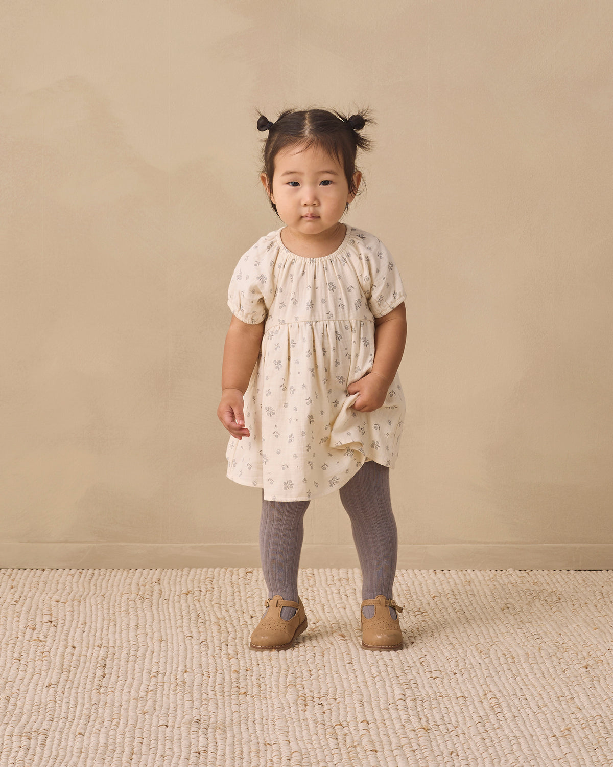 A toddler girl stands in a studio wearing a cream short-sleeve dress with a small beige floral print, gray tights, and tan shoes against a warm beige backdrop.