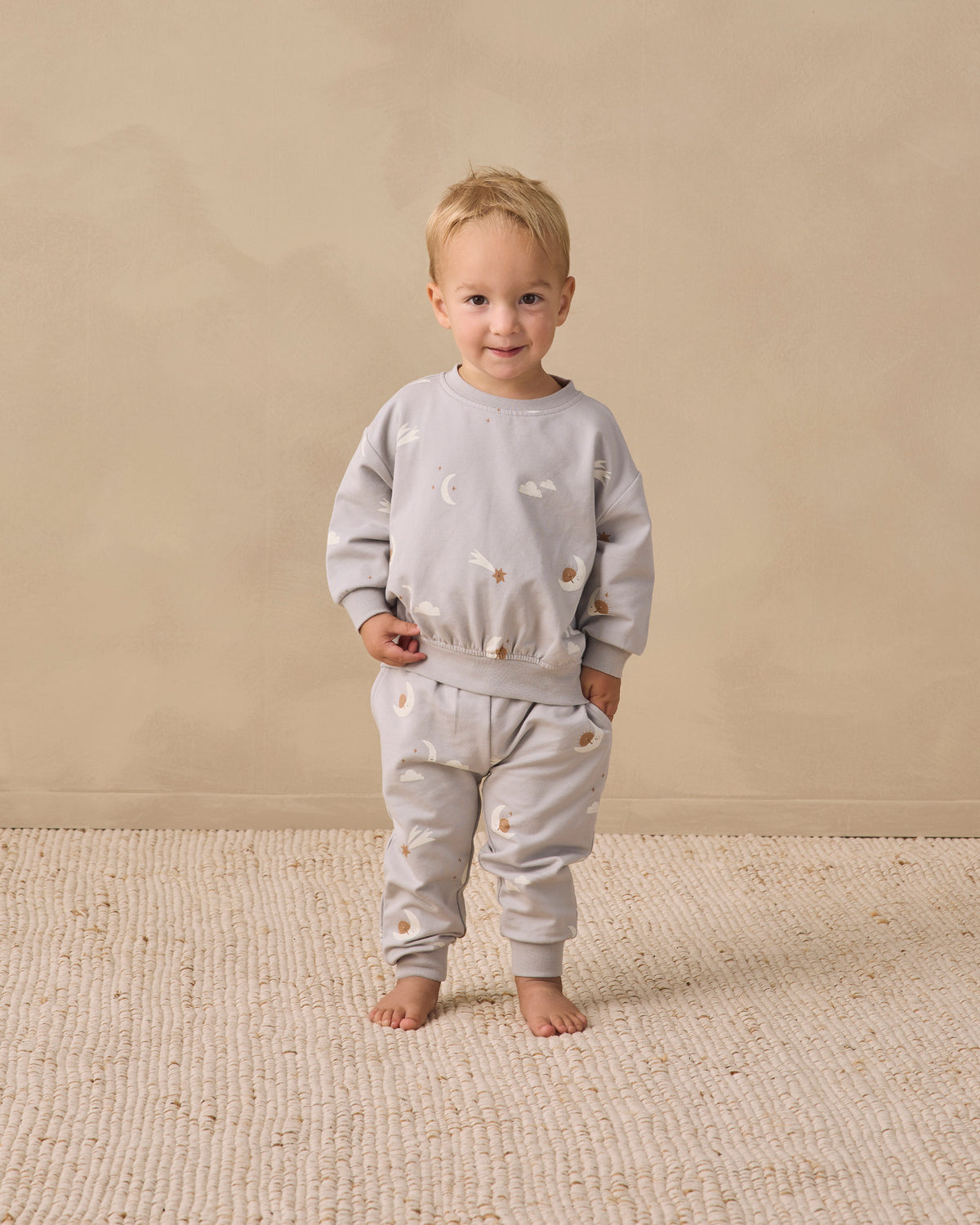 A toddler stands barefoot on a textured cream rug against a warm beige studio wall, wearing a light gray sweatshirt and matching jogger pants with small embroidered motifs.