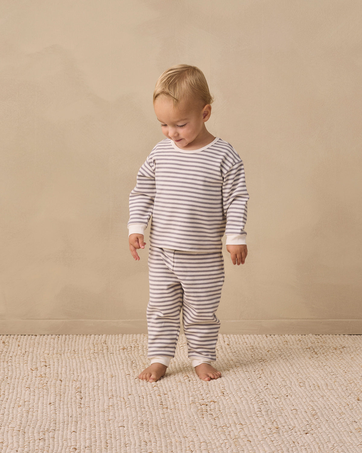 A toddler standing barefoot on a textured cream rug against a warm beige wall, wearing a long-sleeve and matching striped pajama set in beige and white.