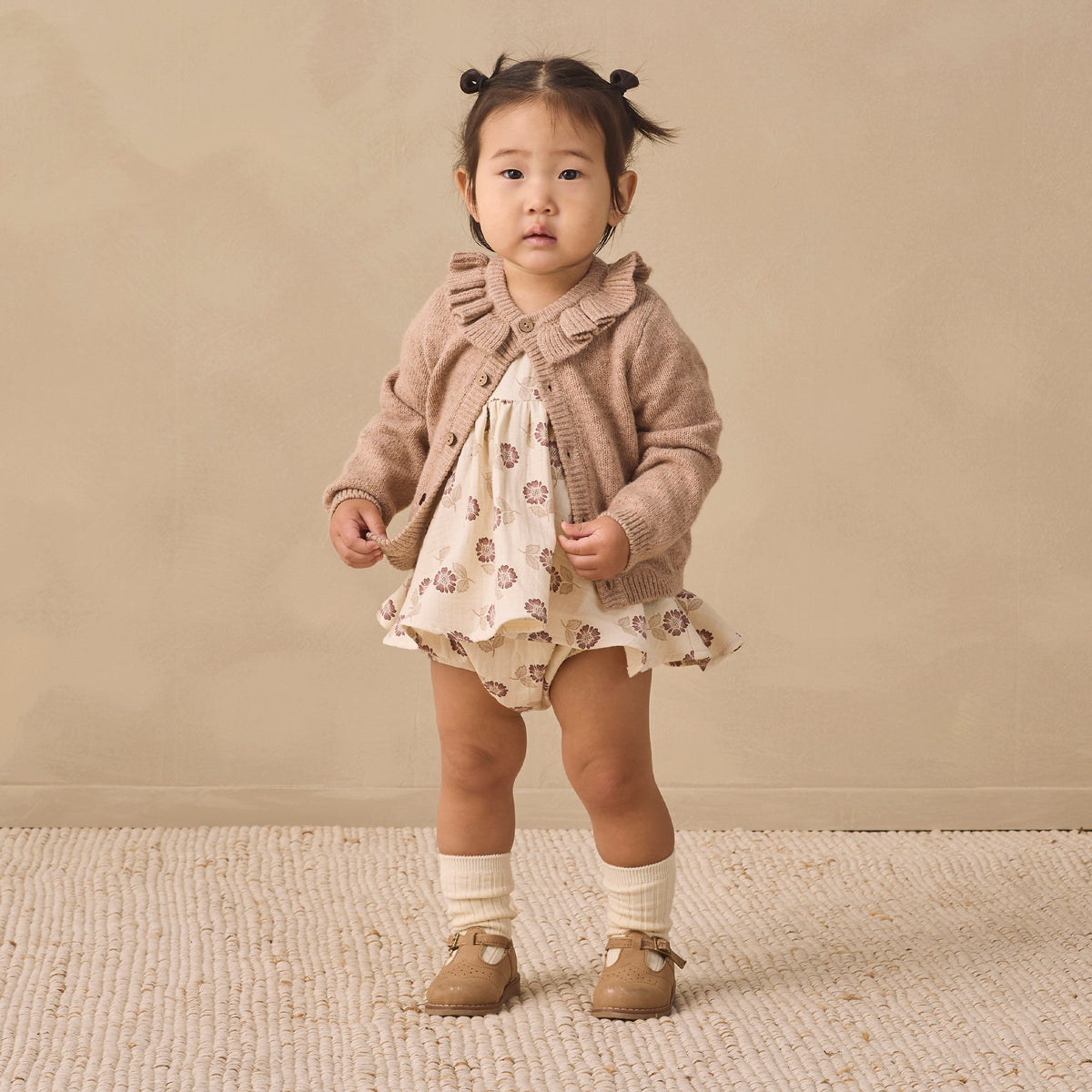 A toddler stands against a warm beige studio backdrop wearing a cream dress with a brown animal print, a taupe ruffled cardigan, white socks, and tan shoes.