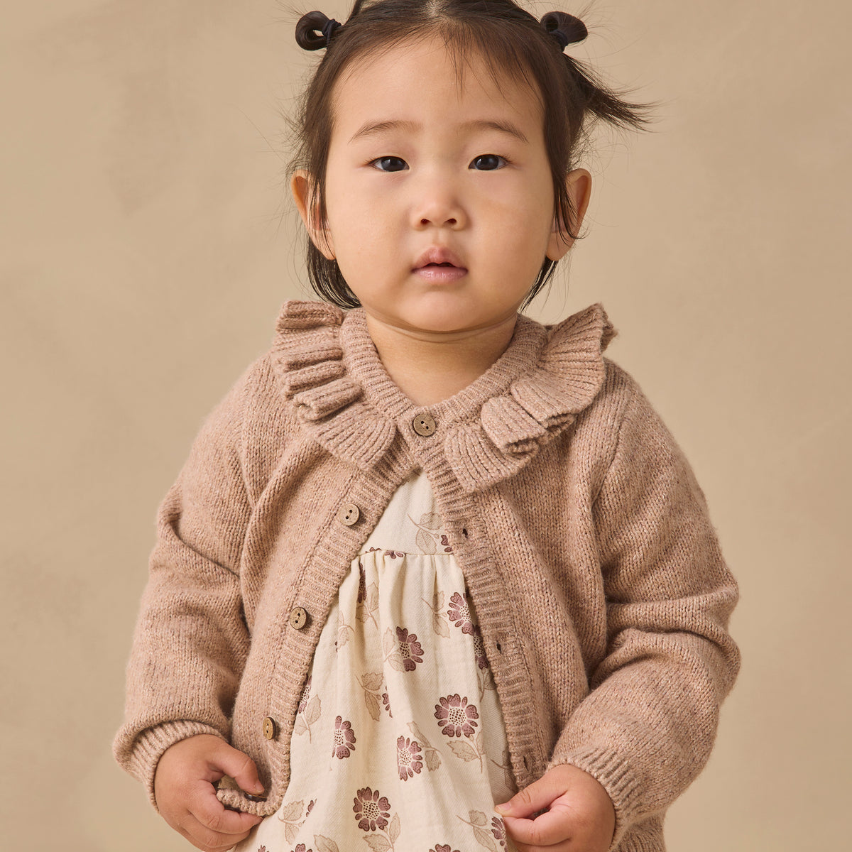A toddler girl with dark hair in two buns wearing a beige knit cardigan with a ruffled collar over a cream dress with a brown floral pattern, standing against a solid beige background.