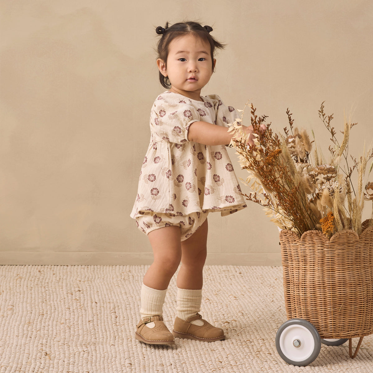 A toddler girl stands in a studio setting wearing a cream-colored short-sleeve romper with a brown floral pattern, paired with cream socks and tan Mary Jane shoes. She is positioned next to a wicker basket filled with dried flowers against a neutral beige background.