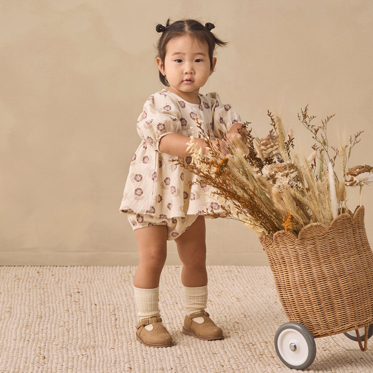 A toddler girl stands in a studio setting next to a wicker cart filled with dried flowers, wearing a cream short-sleeve romper with a brown floral pattern, cream socks, and tan shoes.