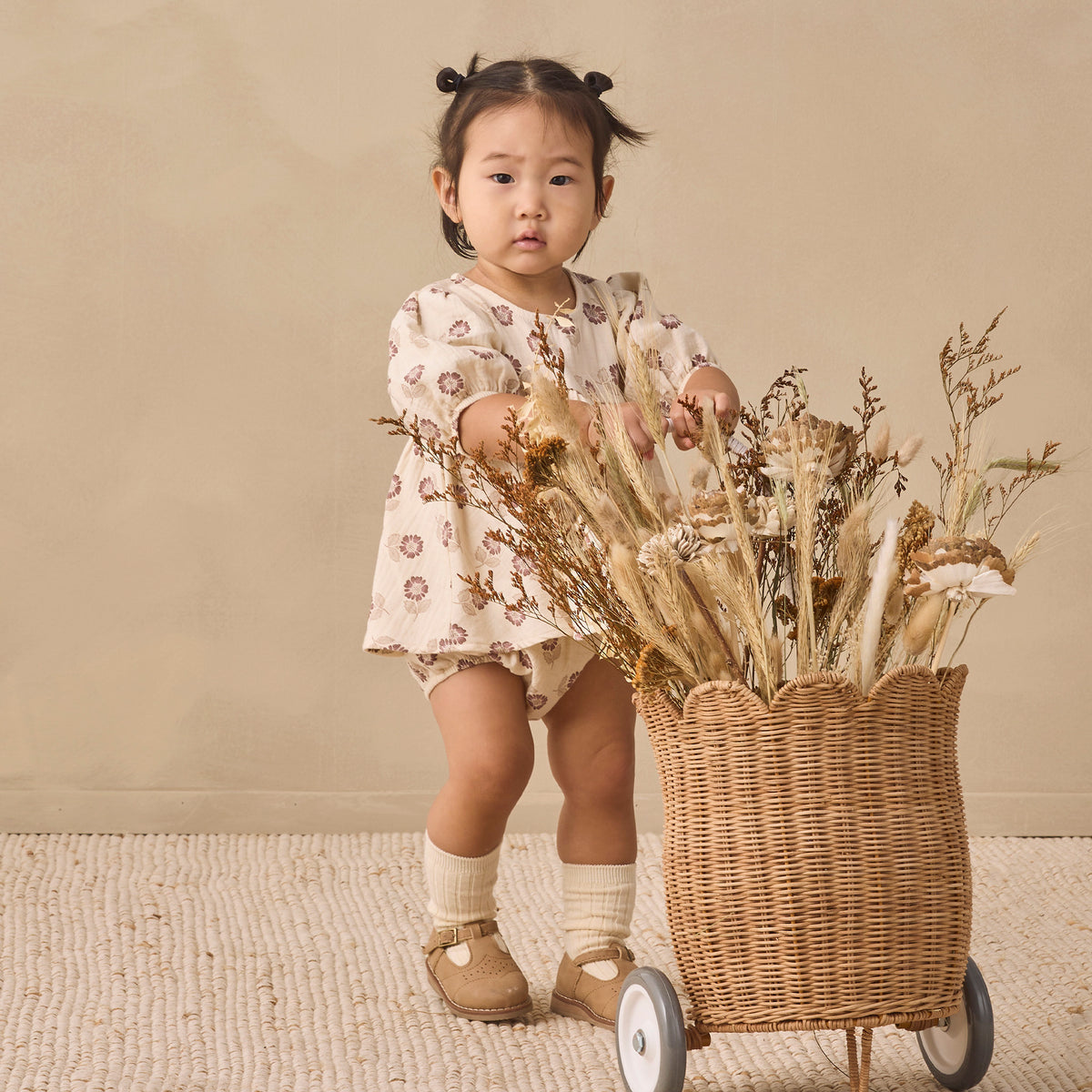 A toddler girl stands behind a wicker cart filled with dried flowers in a studio setting. She is wearing a cream-colored romper with a brown floral pattern, cream socks, and tan shoes, against a warm beige background.
