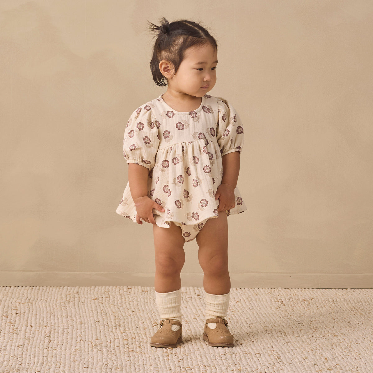 A toddler girl stands on a textured cream rug against a warm beige wall, wearing a cream short-sleeve romper with a brown floral pattern, cream knit socks, and tan shoes.