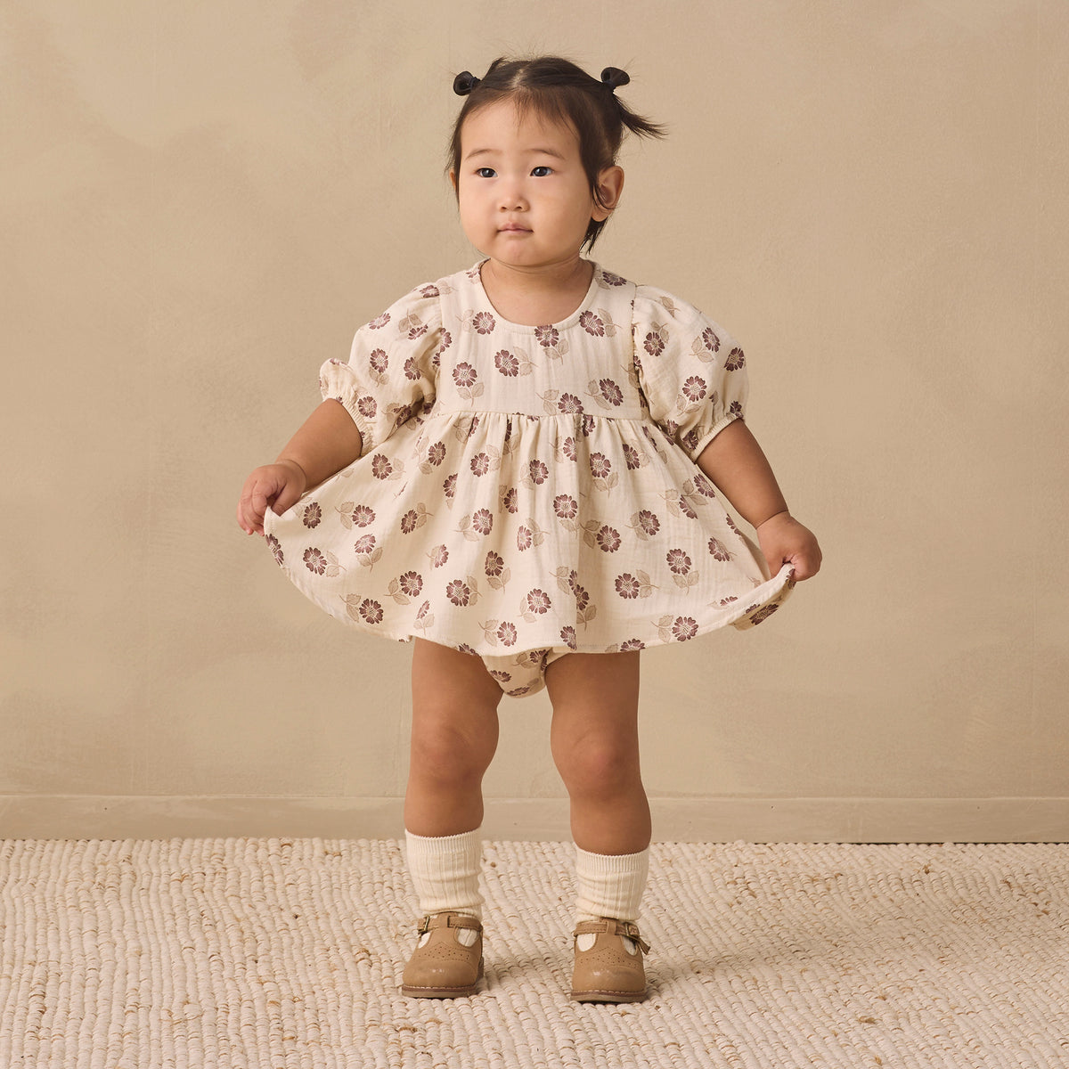 A toddler girl stands in a studio wearing a cream-colored puff-sleeve dress with a brown floral pattern, paired with cream socks and tan shoes, against a warm beige backdrop.