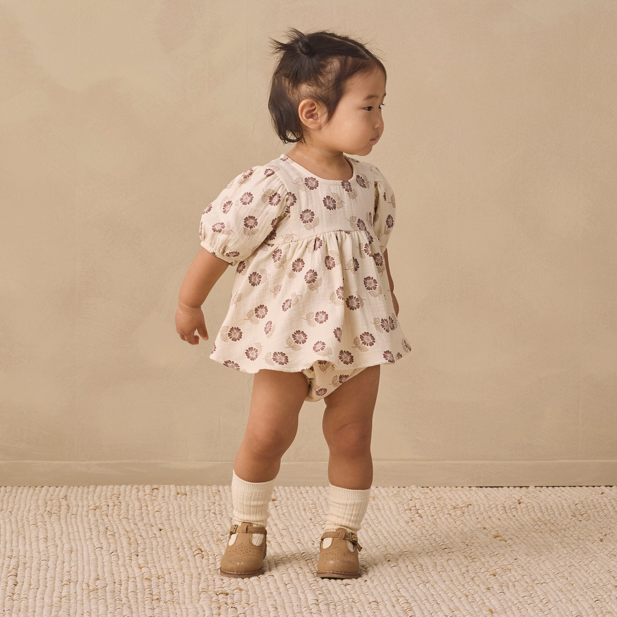 A toddler girl wearing a cream-colored short-sleeve romper with a brown floral pattern, standing on a textured beige rug against a neutral beige background.
