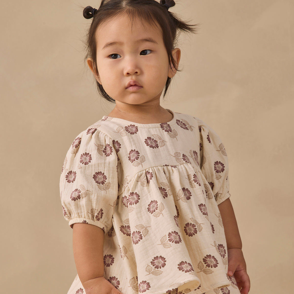 A toddler girl with her hair in two buns wears a cream-colored short-sleeve dress with a brown floral pattern, standing against a solid beige background.