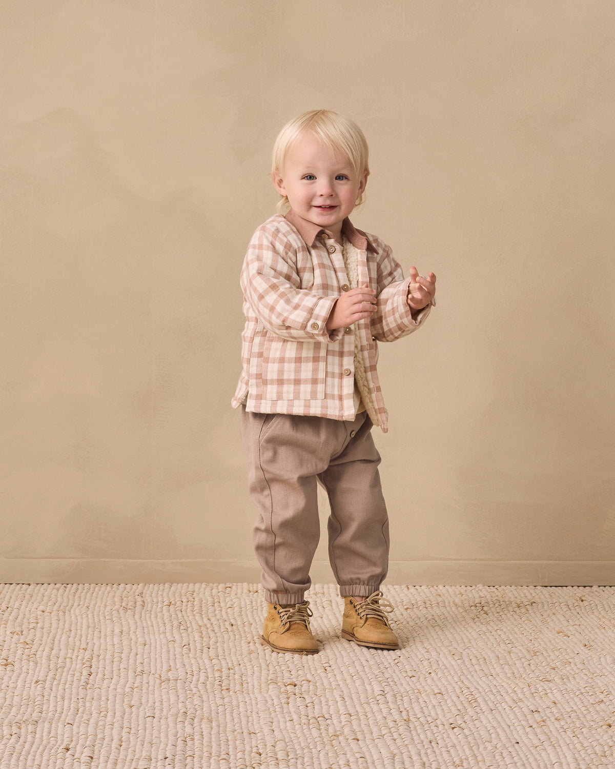 A toddler standing in a studio setting, wearing a beige and brown plaid button-up shirt, taupe pants, and tan boots against a warm beige backdrop.
