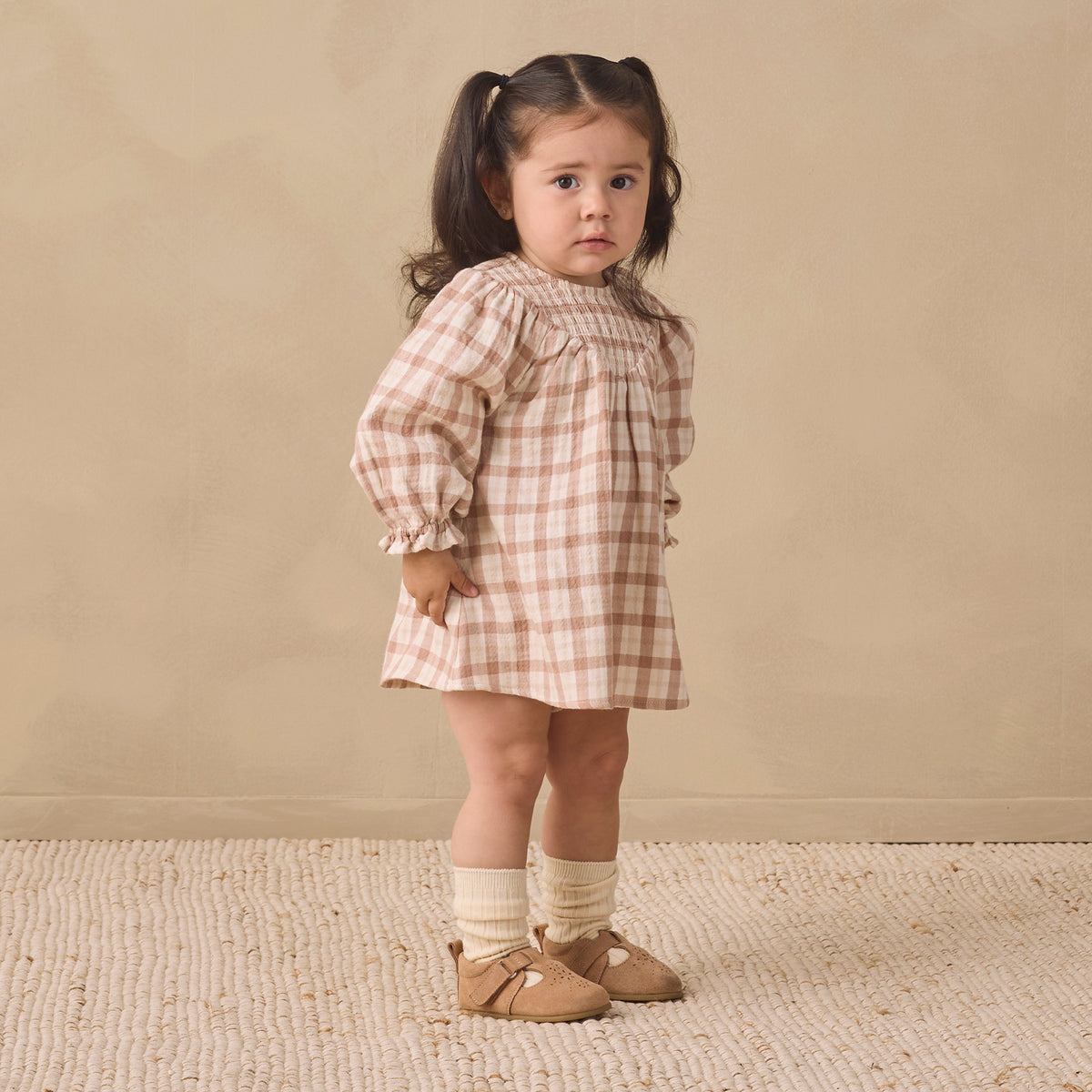 A young girl with dark hair in a ponytail stands in a studio wearing a beige and white gingham dress, cream socks, and tan shoes against a warm beige background.