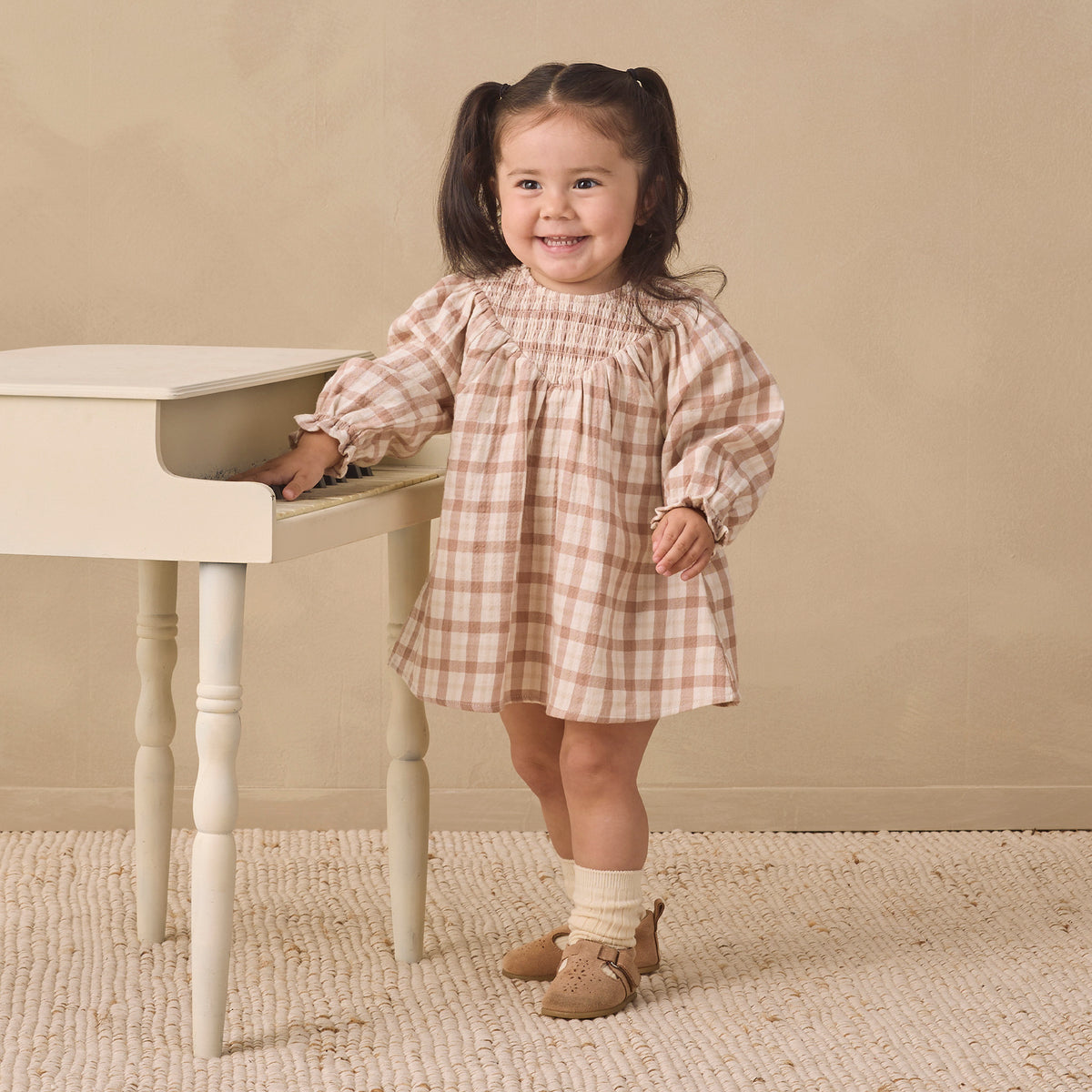 A young girl with dark hair in pigtails stands next to a small white table, wearing a beige and white gingham dress, cream socks, and tan shoes, against a neutral beige background.