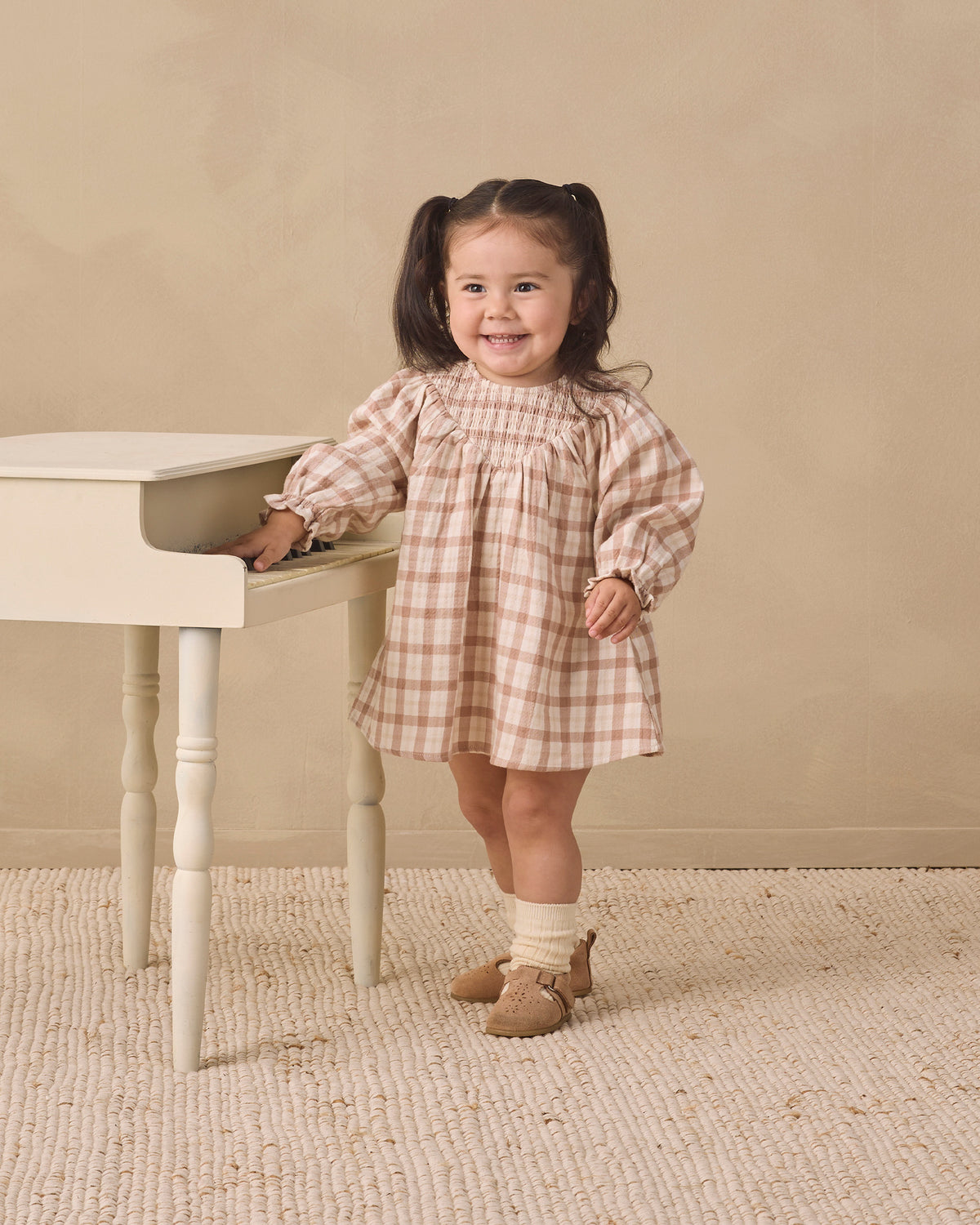 A young girl with dark hair in pigtails stands next to a small white table, wearing a beige and white gingham dress, cream socks, and tan shoes, against a neutral beige background.