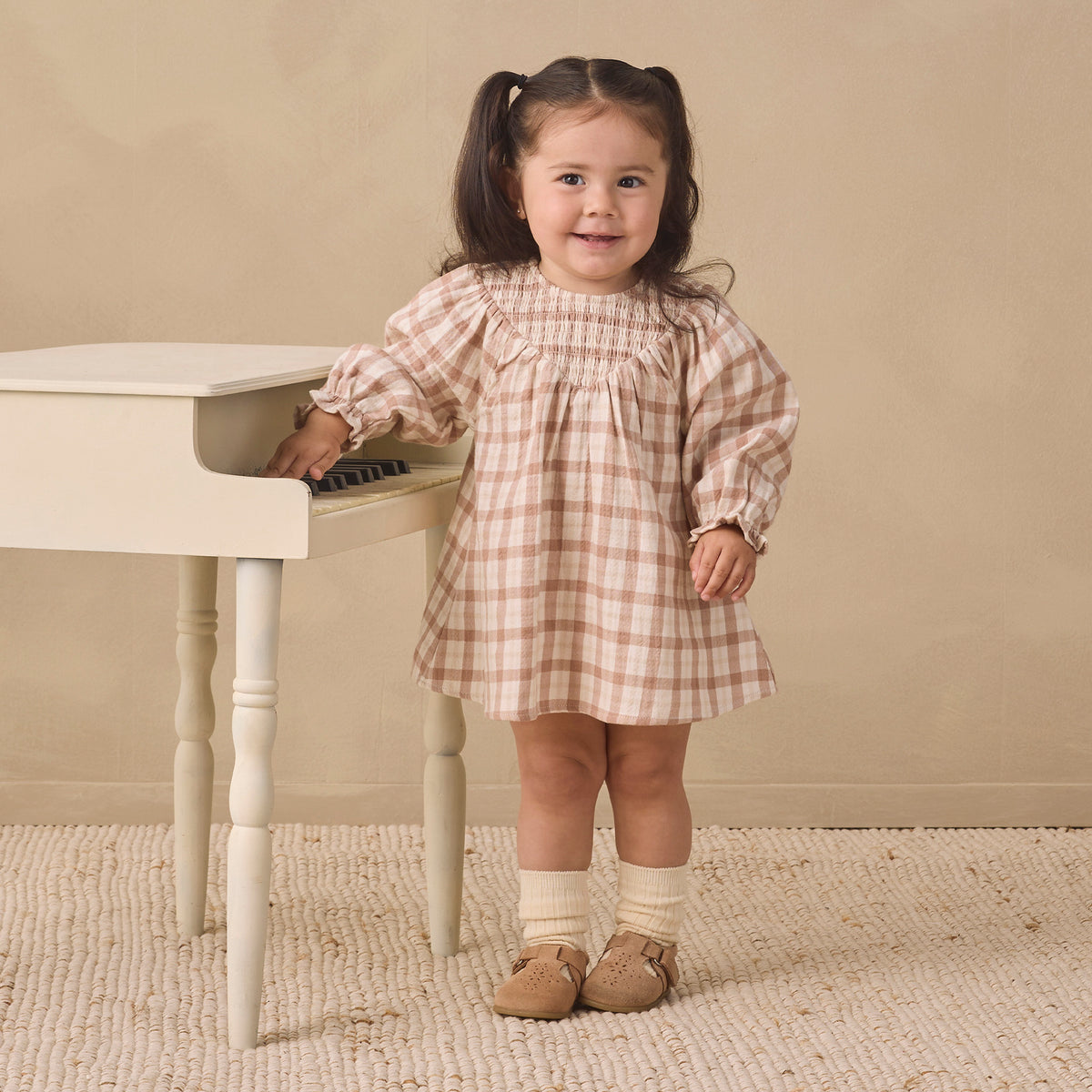 A young girl with dark hair in pigtails stands next to a small white piano, wearing a beige and white gingham dress, cream socks, and tan shoes, set against a neutral beige wall and textured rug.