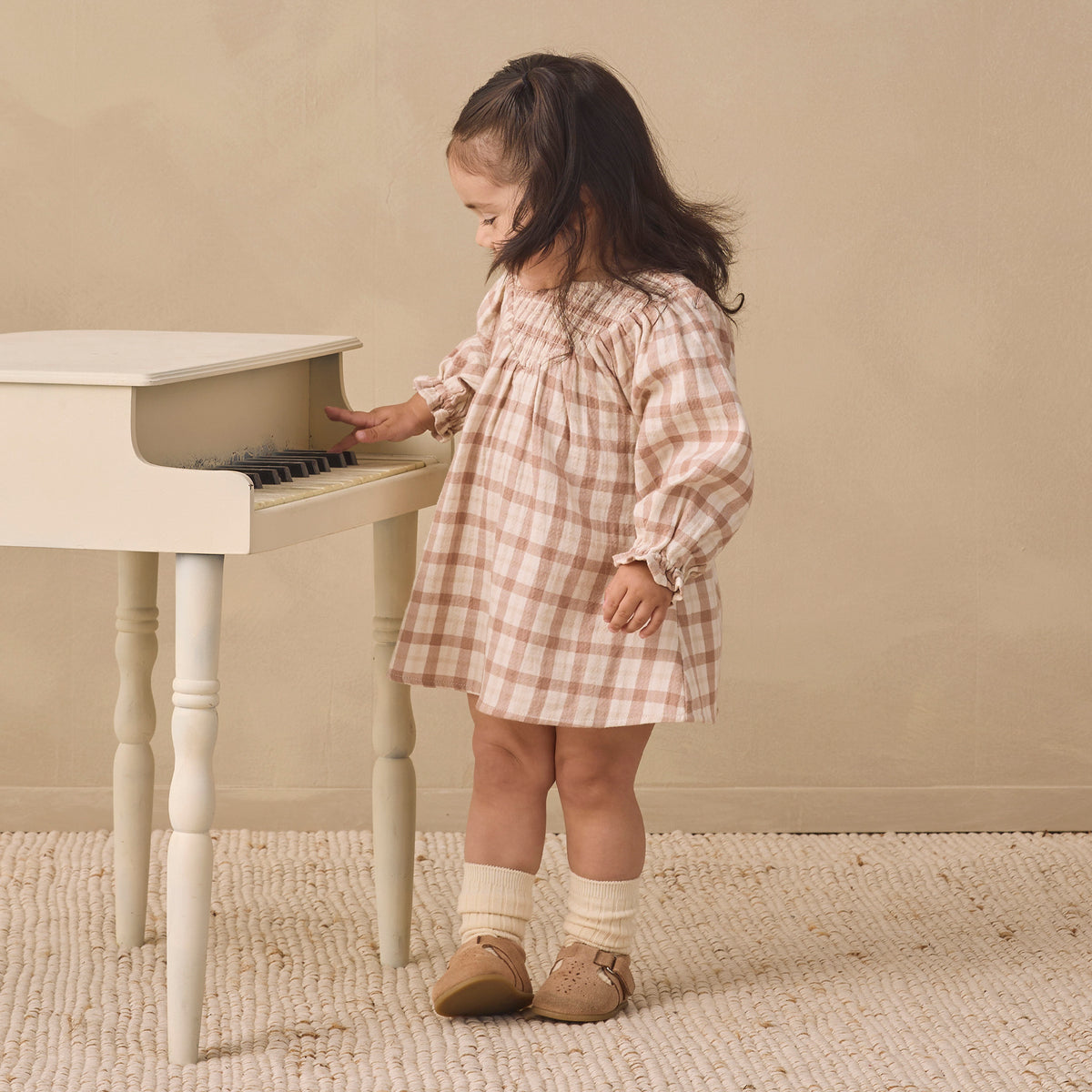 A toddler girl wearing a pink and beige gingham dress stands next to a small white piano, touching the keys. She is wearing cream socks and tan shoes in a warm, neutral-toned room with a textured rug.