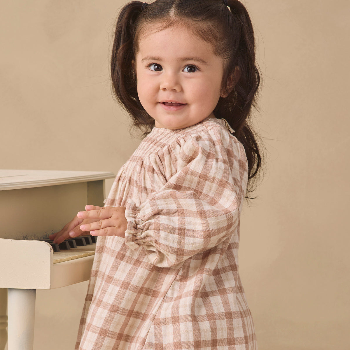 A young girl with dark brown pigtails stands next to a white piano, wearing a beige and brown plaid long-sleeve dress against a solid beige background.