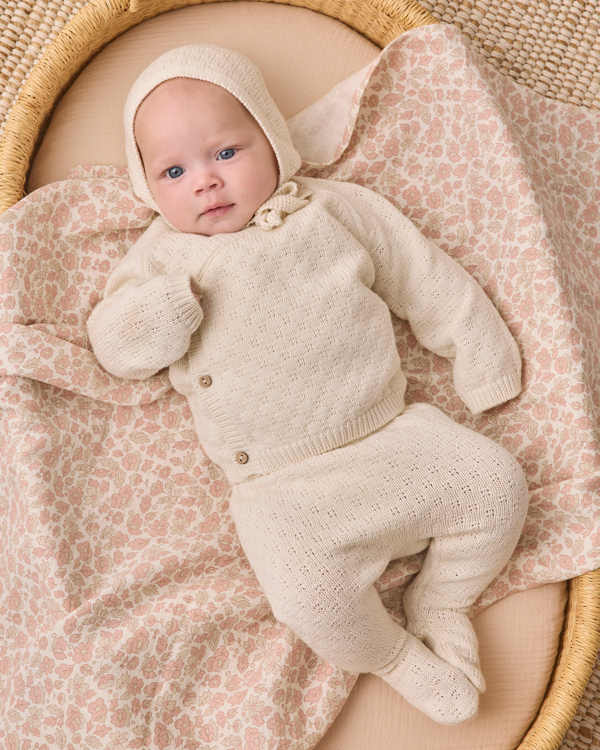 A baby wearing a cream-colored knit bonnet, cardigan, and footie sleeper set, lying on a pink and beige patterned blanket inside a woven bassinet.