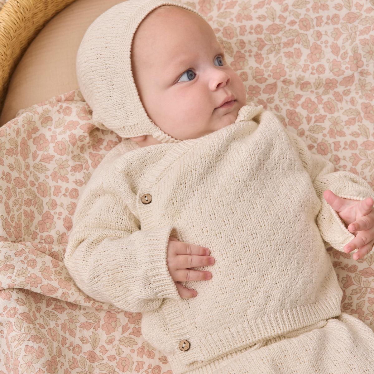 A baby wearing a cream-colored knit bonnet and matching cardigan lies on a patterned blanket inside a woven basket.