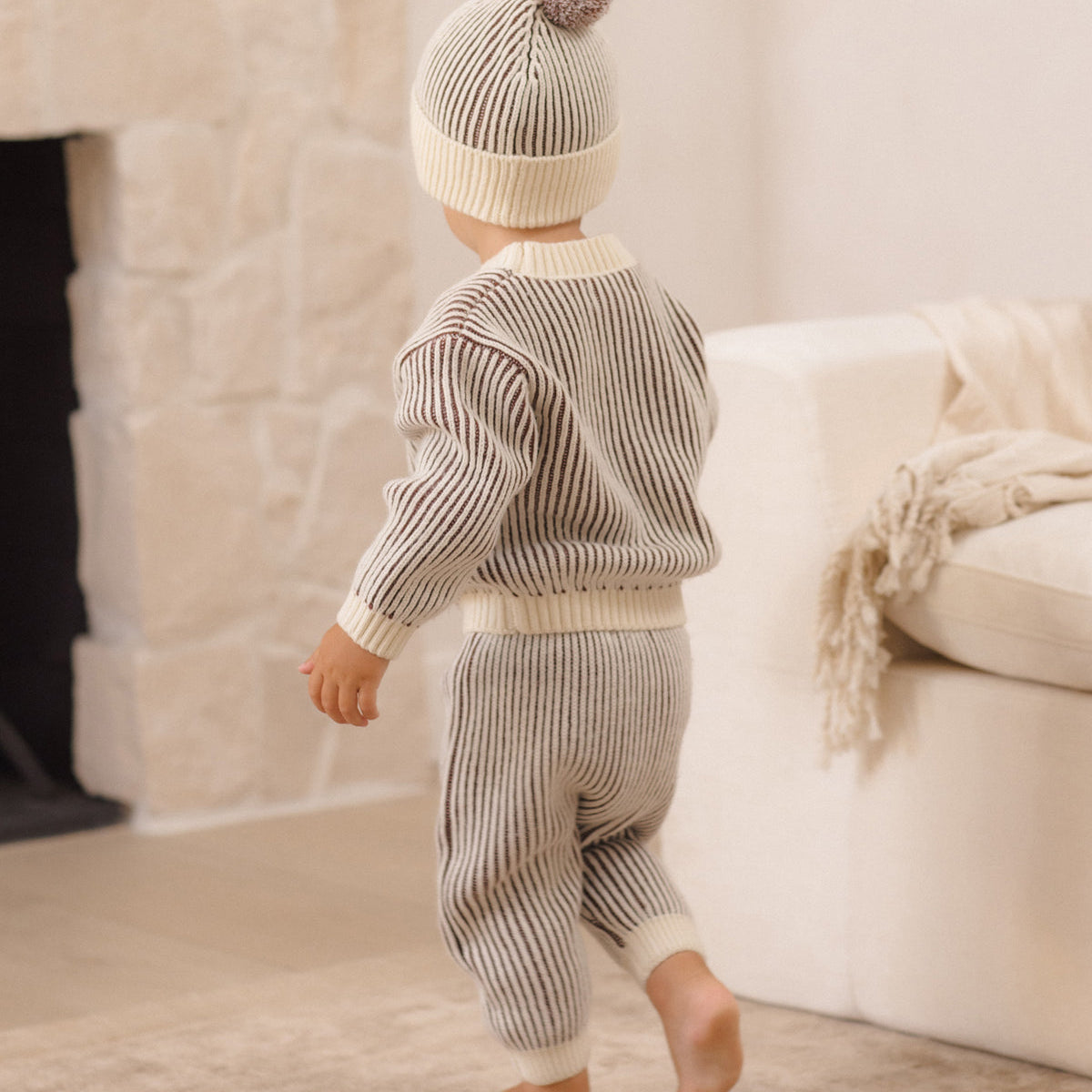A baby wearing a matching ribbed knit two-piece outfit with a pom-pom beanie, shown from behind in a softly lit living room.