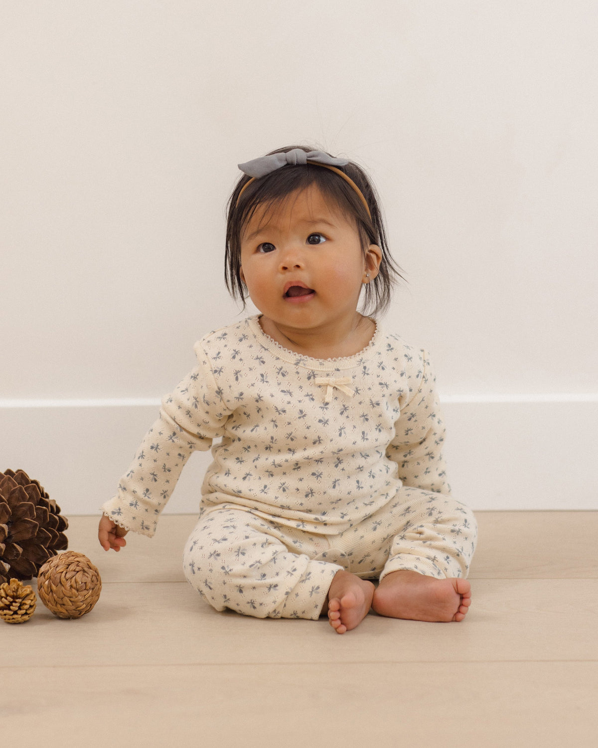 A baby sitting on a light wooden floor against a cream wall, wearing a cream-colored long-sleeve romper with a small blue floral pattern and a matching headband.