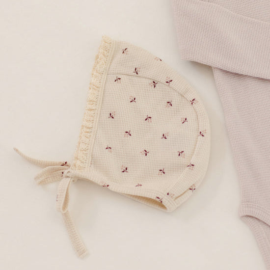 A cream-colored waffle-knit baby bonnet with tiny red embroidered bows and a ruffled trim, styled next to a pale pink waffle-knit baby bodysuit on a warm off-white background.