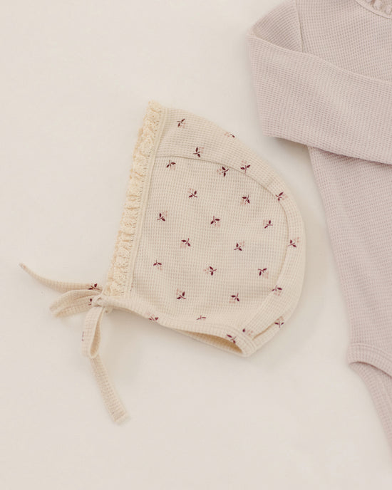 A cream-colored waffle-knit baby bonnet with tiny red embroidered bows and a ruffled trim, styled next to a pale pink waffle-knit baby bodysuit on a warm off-white background.