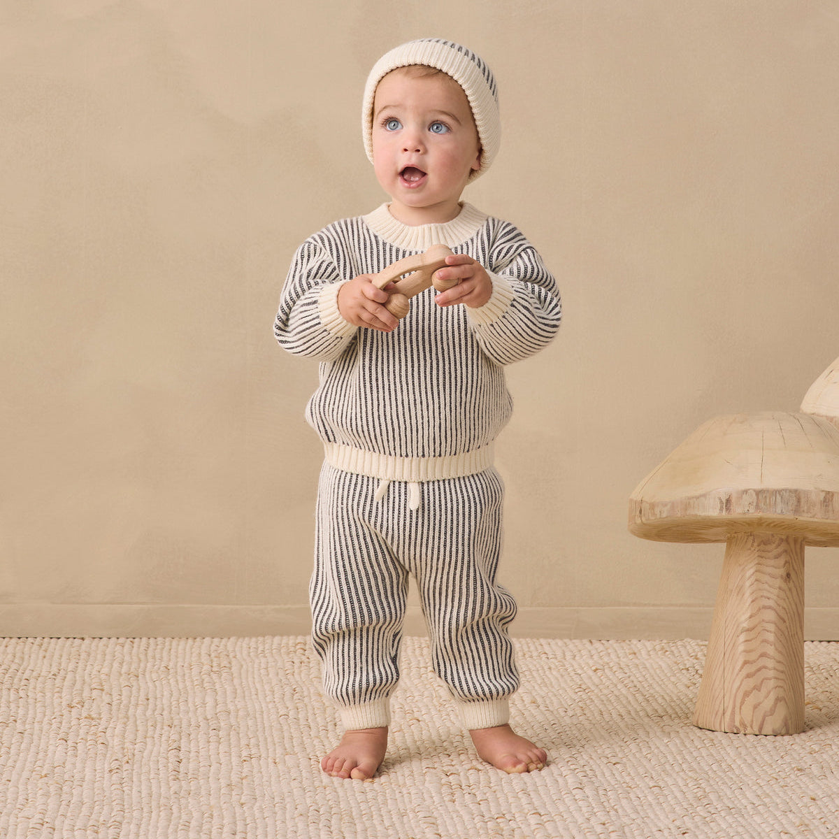 A baby wearing a matching striped knit outfit with a beanie hat, standing barefoot on a textured rug in a warm neutral room. The outfit consists of a long-sleeve top and pants in a gray and white vertical stripe pattern with cream-colored ribbed trim.