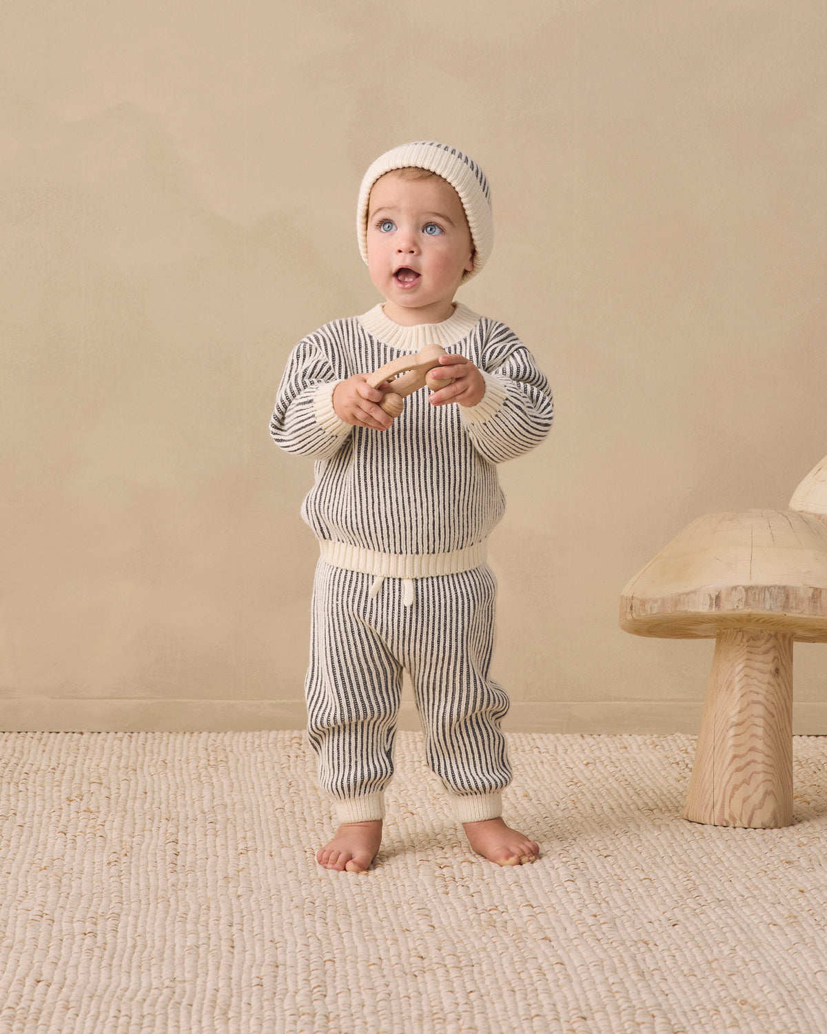 A baby wearing a matching striped knit outfit with a beanie hat, standing barefoot on a textured rug in a warm neutral room. The outfit consists of a long-sleeve top and pants in a gray and white vertical stripe pattern with cream-colored ribbed trim.